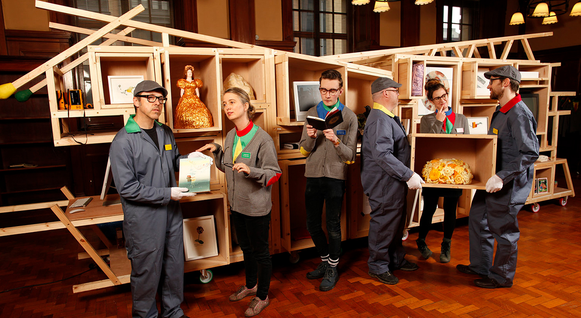 Wooden sculpture with four people standing and holding small art works 